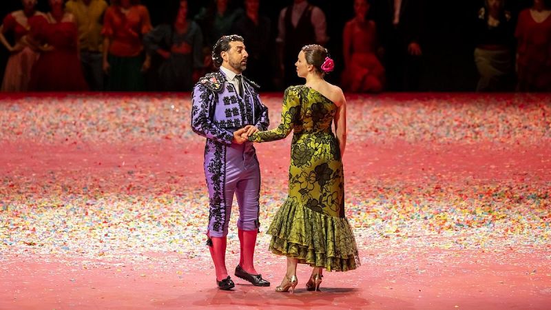 Dos protagonistas de una ópera se miran con complicidad sobre una tarima con confeti, él con traje de luces lavanda y ella con vestido verde oliva.