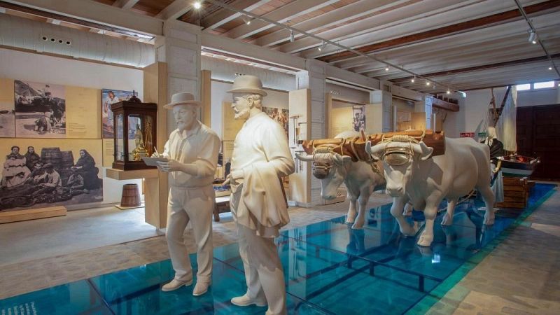 Instalación escultórica en una sala de exposiciones que incluye figuras humanas de época, bueyes y una barca sobre un suelo azul translúcido.