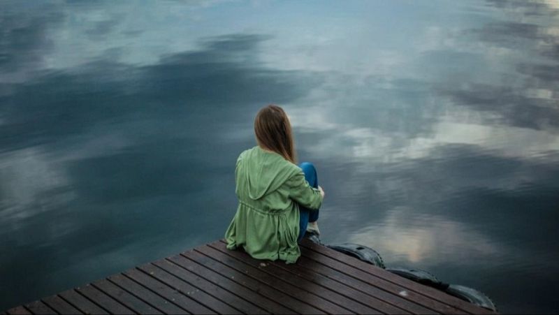 Persona joven de espaldas sentada en un muelle de madera frente al agua tranquila bajo un cielo nublado.