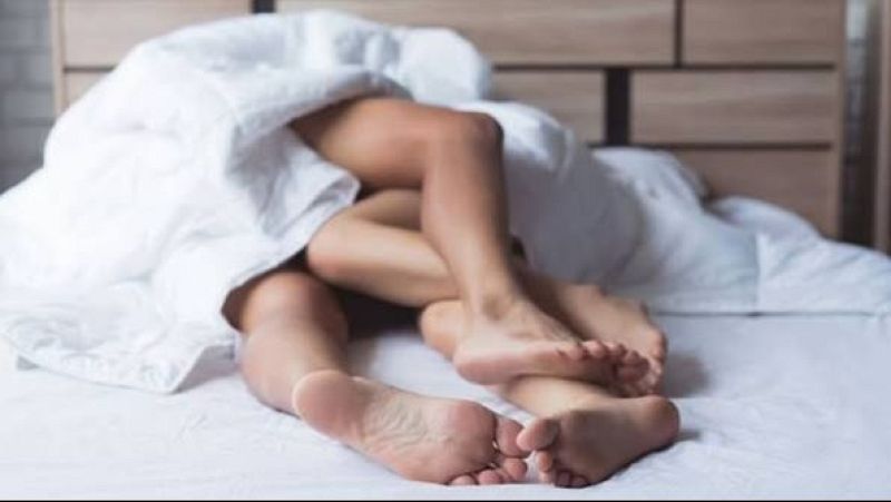 Plano medio-corto de una cama blanca con las piernas entrelazadas de dos personas y un fondo desenfocado.