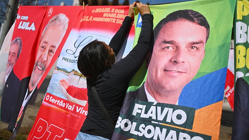 Mujer de espaldas arreglando toallas de merchandising político brasileño de Lula da Silva y Flávio Bolsonaro en un puesto callejero.