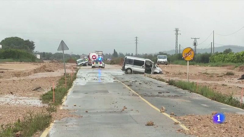 Furgoneta gris atrapada en el lodo de una carretera secundaria afectada por lluvias, con veh�culos de emergencia al fondo bajo un cielo gris.