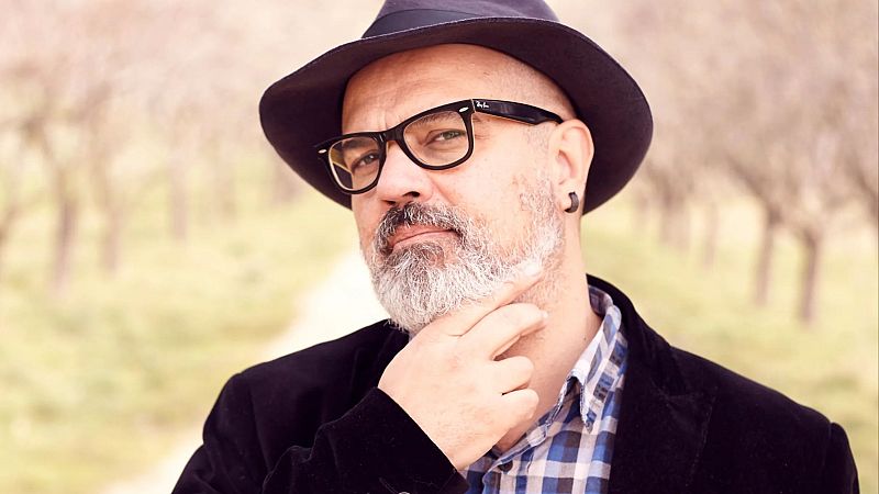Plano medio corto del músico Leo Minax al aire libre, con sombrero negro, gafas de pasta y una pose reflexiva.