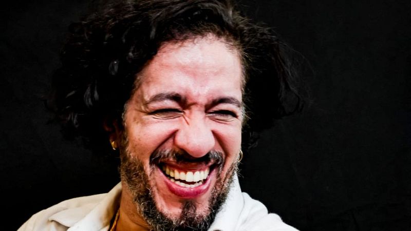 Primer plano de Jean Wyllys riendo con los ojos cerrados, luciendo cabello rizado, barba, aros dorados y camisa blanca sobre fondo oscuro.