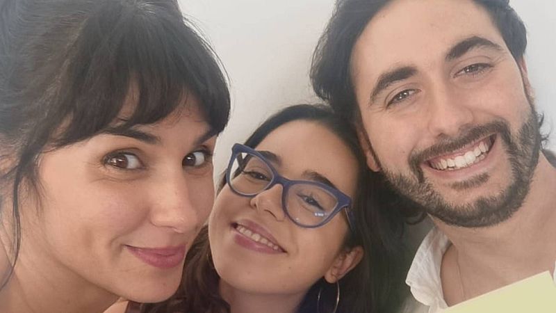 Selfi de tres jóvenes sonrientes con fondo blanco: una mujer con pelo recogido, otra con gafas y un hombre con barba y camisa blanca.