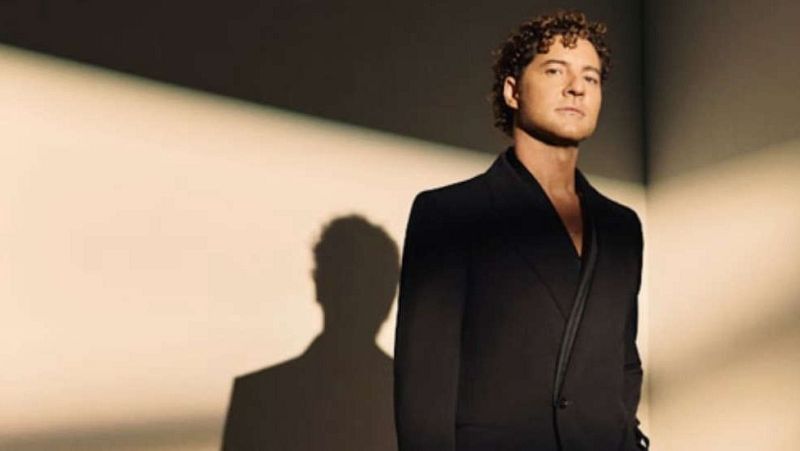 Plano medio del cantante David Bisbal vistiendo chaqueta negra sobre fondo beige, con su sombra proyectada en la pared.