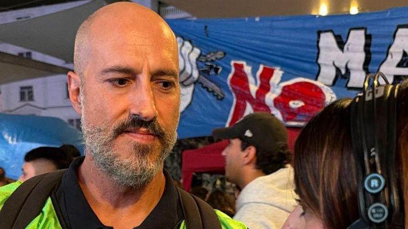 El político Christian Castillo aparece serio en una manifestación al aire libre con tirantes reflectantes, frente a una pancarta con la palabra "No".
