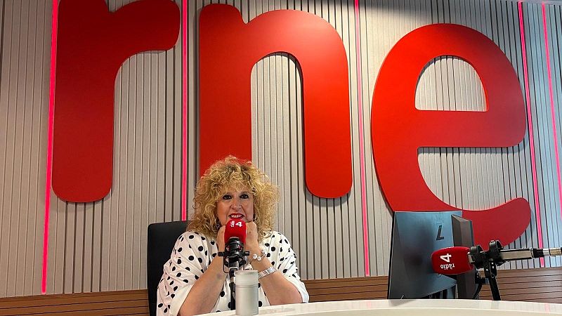 La periodista S�lvia Tarragona en un estudio de radio, sentada ante una mesa con micr�fonos con capuch�n rojo y fondo de paneles ac�sticos con el logotipo de rne.