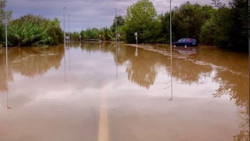 Carretera completamente inundada con agua marr�n y un coche azul atrapado en la orilla, rodeada de �rboles y se�ales de tr�fico parcialment ocultas.
