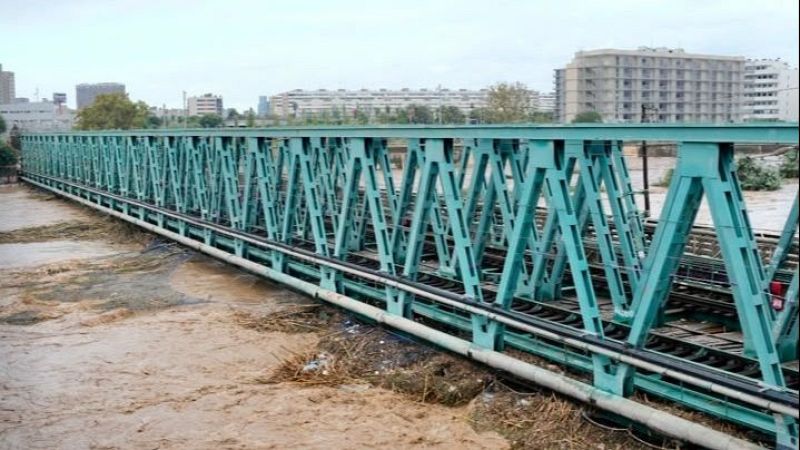Puente ferroviario met�lico verde sobre un r�o crecido y fangoso por lluvias, con edificios urbanos al fondo bajo un cielo gris.