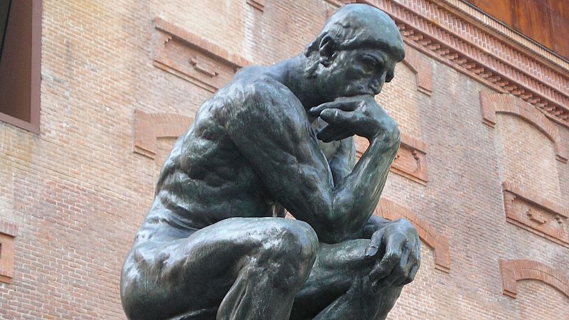 Escultura de bronce 'El pensador' de Rodin, que representa a un hombre sentado y pensativo frente a la fachada de ladrillo de un edificio.