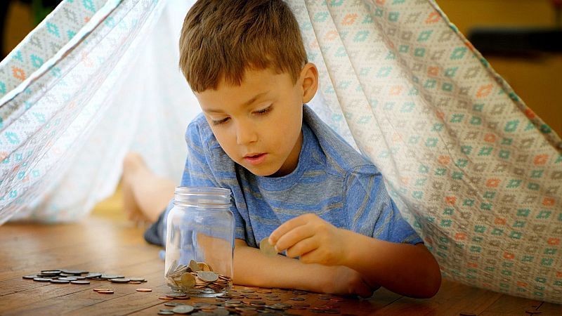 Un niño en camiseta azul recostado en un suelo de madera interactúa con un tarro de cristal lleno de monedas dentro de una tienda de campaña.