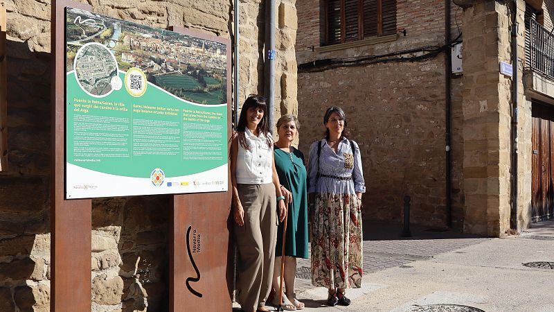 Tres mujeres posan junto a un panel turístico en una calle empedrada, flanqueadas por edificios rústicos de piedra.