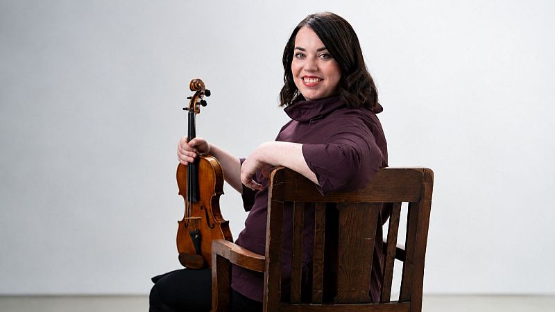 Retrato de media distancia de una mujer con cabello castaño y blusa morada, sentada en una silla de madera mientras sostiene un violín en un estudio.