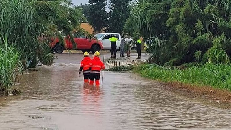 Dos bomberos con trajes de rescate naranjas caminan con el agua hasta los muslos por un camino rural inundado.