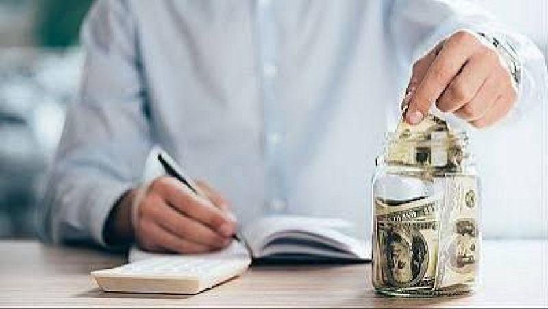 Persona sentada ante un escritorio anotando en una libreta junto a una calculadora y metiendo un billete de cien d�lares en un tarro de cristal que hace de hucha.