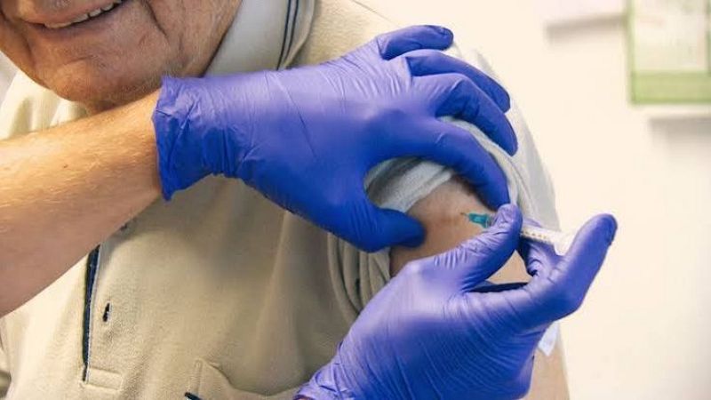 Un hombre mayor sonr�e mientras recibe una inyecci�n en el brazo por parte de un sanitario con guantes azules en una consulta m�dica.