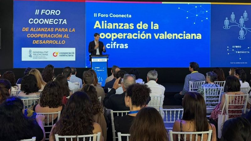 Ponente en un escenario ante una gran pantalla azul con gráficos y textos institucionales, dirigiendo su discurso a una audiencia sentada de espaldas.