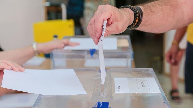 Mano de un votante introduciendo un sobre blanco en una urna electoral transparente de metacrilato con precintos azules.