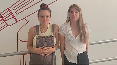 Dos m�sicas posan juntas en plano medio, vistiendo ropa casual en un estudio con fondo blanco y una barandilla met�lica.