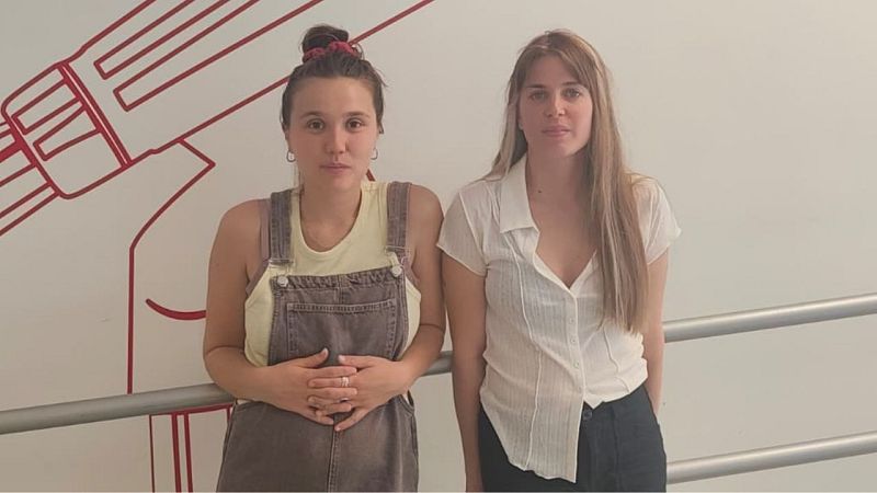Dos músicas posan juntas en plano medio, vistiendo ropa casual en un estudio con fondo blanco y una barandilla metálica.