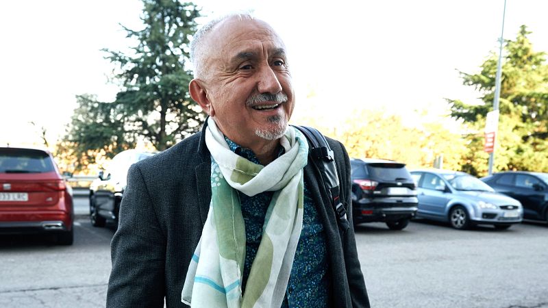 El periodista Josep Cuní camina sonriente en exteriores con americana, fular y mochila, en un entorno de aparcamiento al aire libre.
