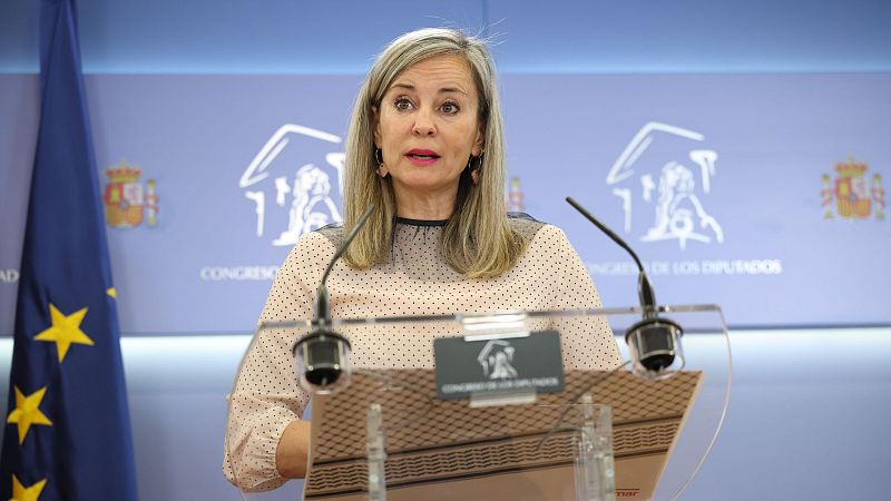 Plano medio de la política Verónica Martínez Barbero en rueda de prensa tras un atril transparente en el Congreso de los Diputados, con panel corporativo azul y bandera europea al fondo.