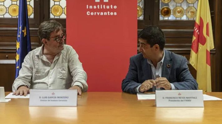 Dos hombres conversan sentados ante una mesa de madera con carteles identificativos, frente a un panel rojo y banderas institucionales.