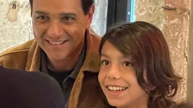 Dos personas sonrientes en un interior r�stico con paredes de piedra, incluyendo al actor Ralph Macchio y un ni�o de pelo largo.