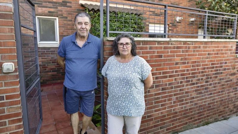 Un hombre y una mujer posan seriamente en la entrada de una vivienda exterior, junto a una cancela met�lica y un muro de ladrillo.