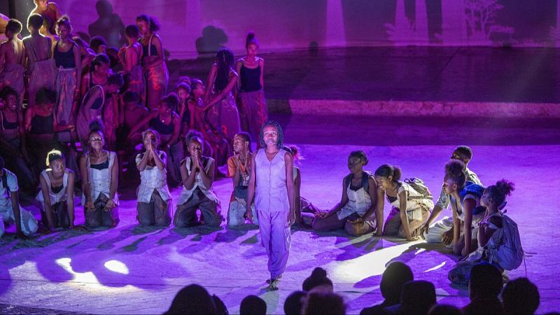 Representación teatral en un escenario con luces violáceas donde una joven intérprete camina al frente, rodeada de actores en el suelo y un coro, bajo la mirada del público.