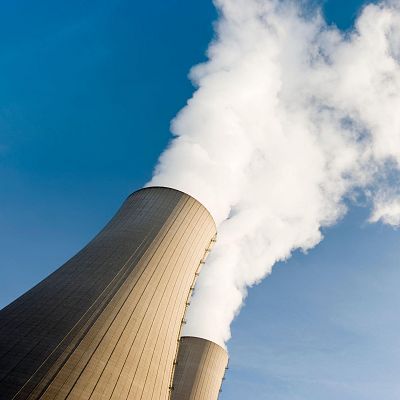 Dos torres de refrigeración grises, con forma cónica, liberan vapor blanco hacia un cielo azul claro. La imagen, tomada desde abajo, destaca la magnitud de las estructuras.