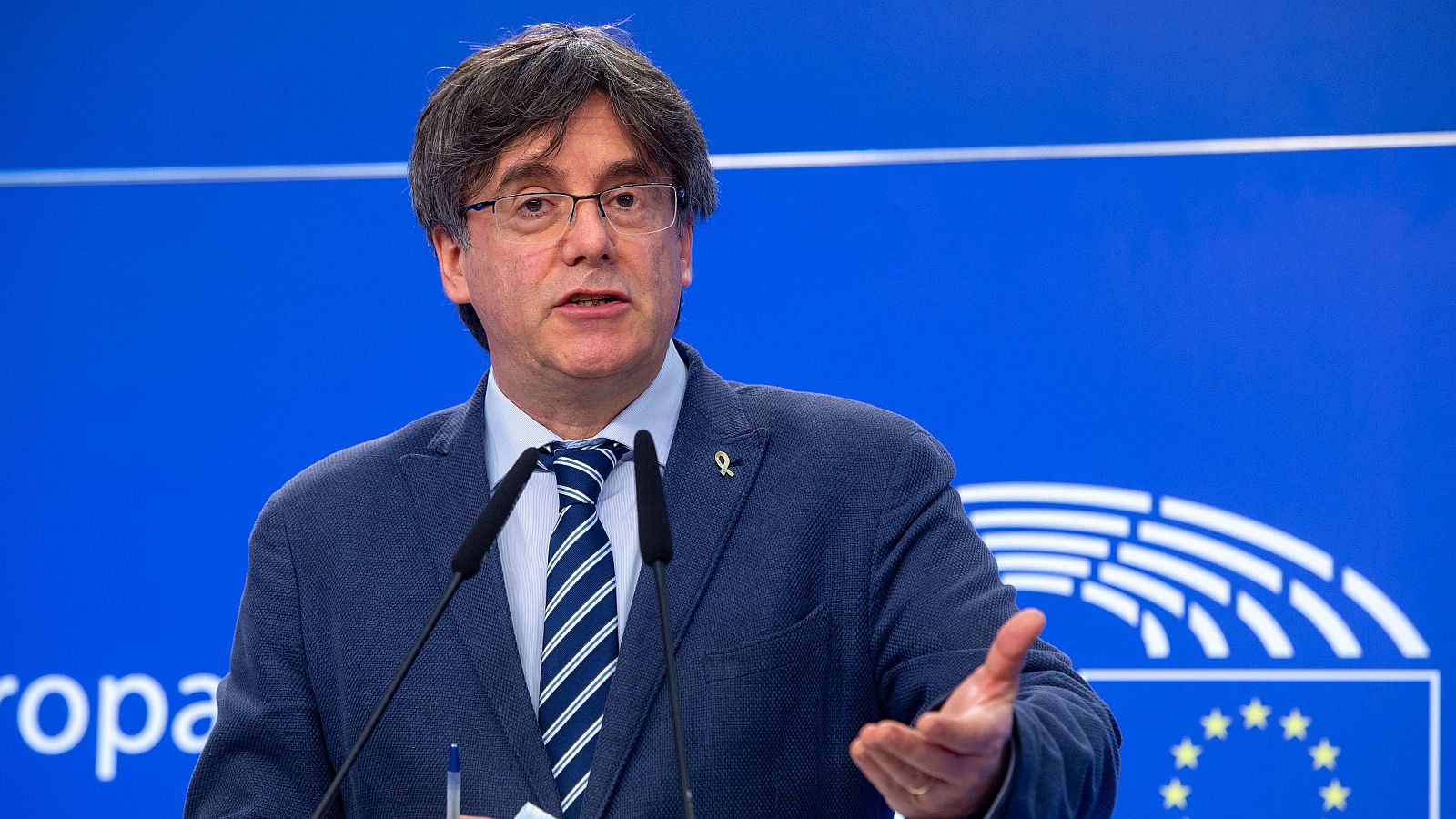 Plano medio del político Carles Puigdemont hablando ante un atril con micrófonos y un lazo amarillo en la solapa, frente a un panel azul del Parlamento Europeo.