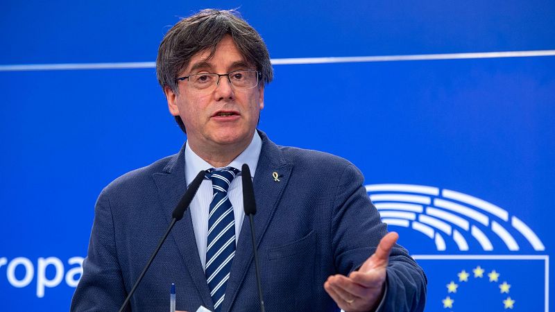 Plano medio del político Carles Puigdemont hablando ante un atril con micrófonos y un lazo amarillo en la solapa, frente a un panel azul del Parlamento Europeo.