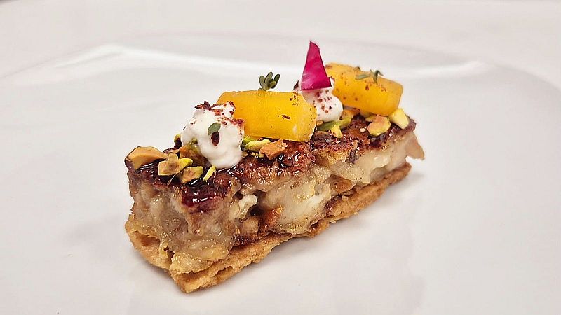 Pintxo de diseño emplatado sobre fondo blanco, con base crujiente, cuerpo caramelizado, dados amarillos, crema blanca, frutos secos y un pétalo magenta.