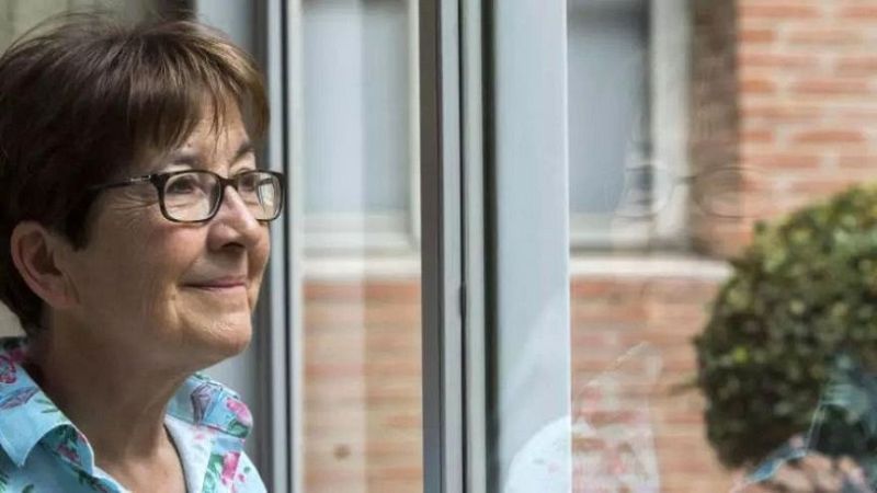 La pedagoga Roser Batlle, con gafas de pasta y camisa estampada, mira sonriente hacia una ventana donde se refleja su rostro.