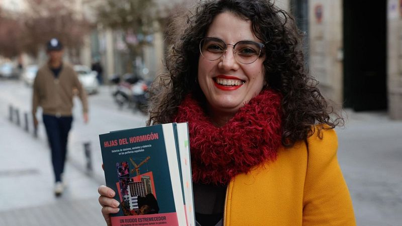 La escritora Aida dos Santos sonríe a la cámara sosteniendo su libro «Hijas del hormigón», vistiendo abrigo mostaza y bufanda granate.