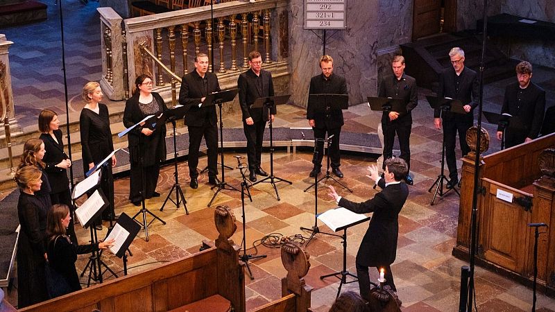Vista picada de un coro mixto de c�mara y su director actuando en una iglesia, con cantantes vestidos de negro frente a atriles y micr�fonos.