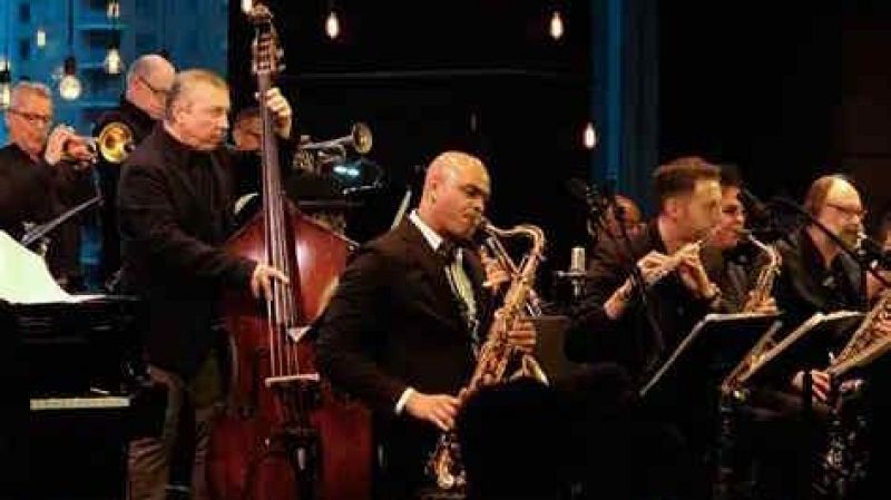 Banda de jazz actuando en directo sobre un escenario con iluminaci�n c�lida y bombillas vintage, destacando un saxofonista solista en el centro y otros m�sicos con contrabajo y vientos.