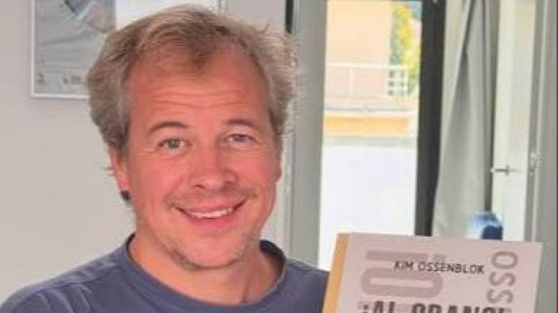 Retrato de medio plano de Kim Ossenblok sonriendo a la cámara, sosteniendo su libro "¡AL GRANO!" en la mano.
