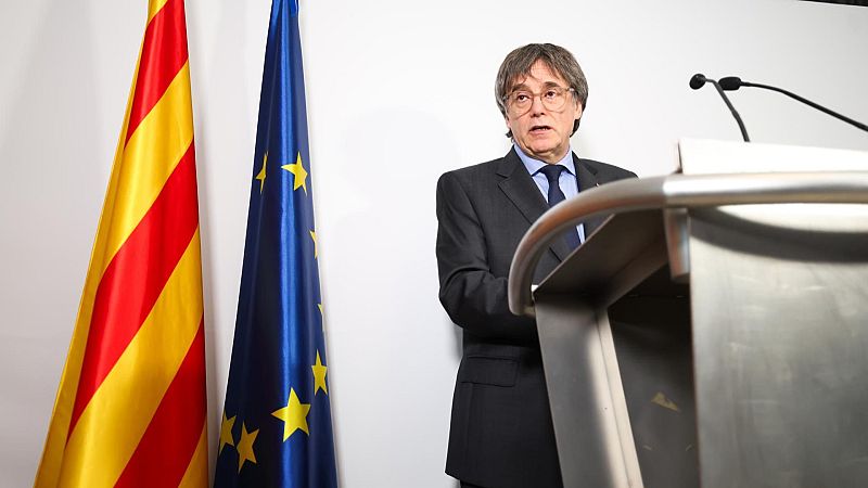 Carles Puigdemont interviene serio ante un atril con traje oscuro, acompañado por las banderas catalana y europea sobre fondo blanco.