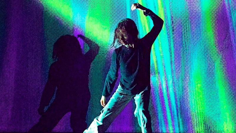 Bailarín en postura expresiva sobre un escenario iluminado con franjas verticales de luz verde, cian y violeta, acompañado por una silueta en sombras.