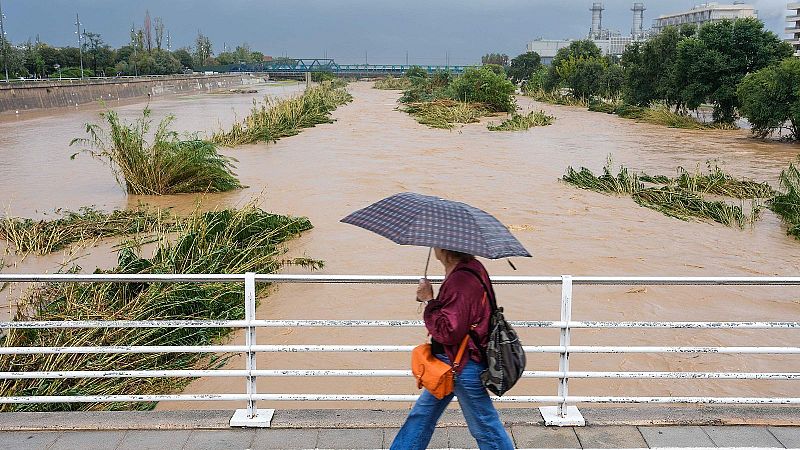 Una persona con paraguas cruza un puente junto a un r�o desbordado de caudal turbio y marr�n bajo un cielo gris de temporal.