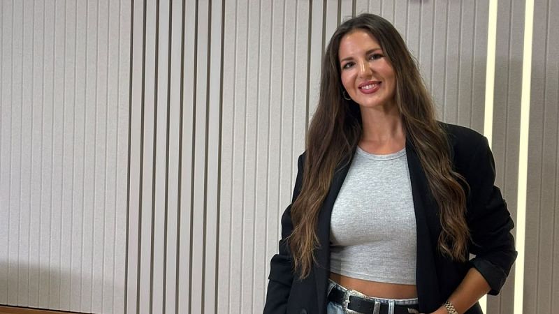 Plano medio de la periodista Georgina Badenas sonriendo, con top gris, blazer negra y vaqueros, frente a una pared de listones de madera y luz tenue.