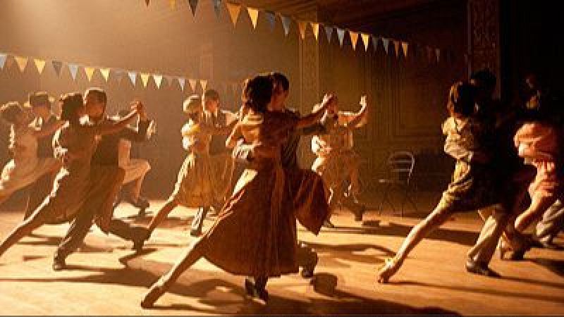 Parejas bailan tango en un sal�n interior con luces doradas y banderines de colores. La mujer viste un vestido ocre y el hombre un traje oscuro.