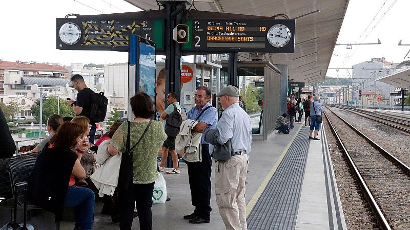 And�n cubierto de una estaci�n de tren con pasajeros esperando junto a las v�as, un panel electr�nico informativo y relojes anal�gicos en la parte superior.