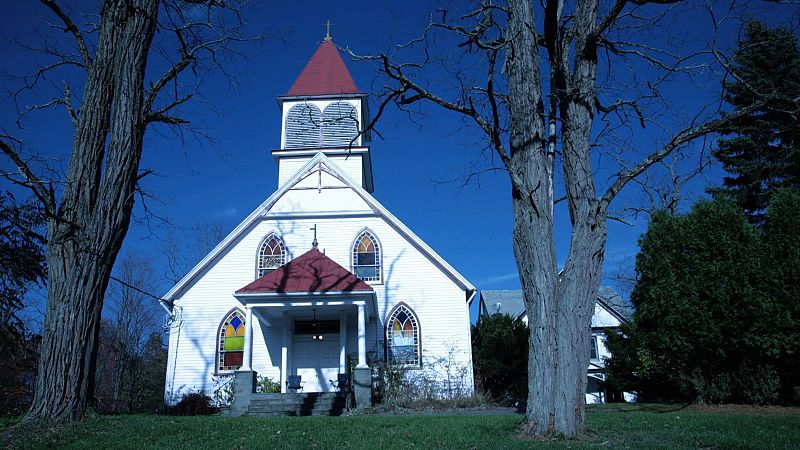 Peque�a iglesia tradicional de madera blanca con campanario rojo y ventanales ojivales, enmarcada por �rboles sobre una colina verde.