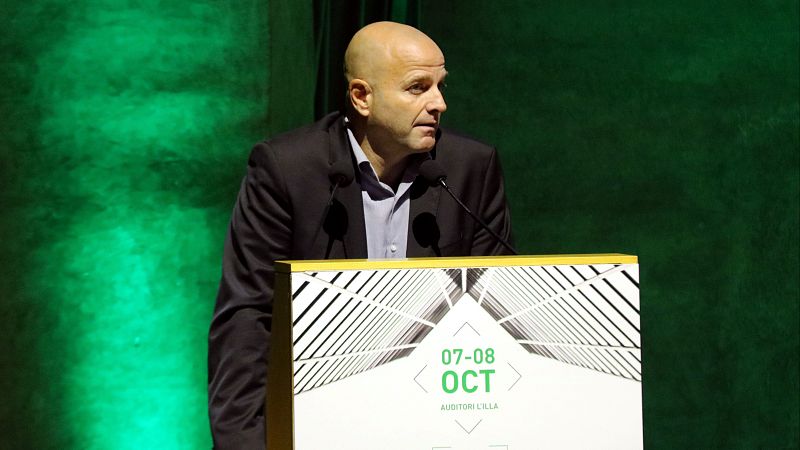 Xavier Vilajoana interviene en un evento ante un atril blanco con gráficos arquitectónicos y fondo verde esmeralda.