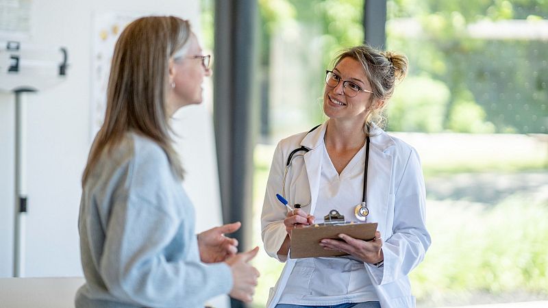 Médica sonriente con bata blanca y estetoscopio escucha atentamente a una paciente en un consultorio luminoso.