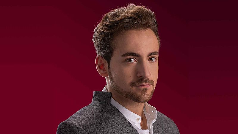 Retrato de estudio de un joven con barba recortada, chaqueta gris y camisa blanca sobre un fondo rojo burdeos.